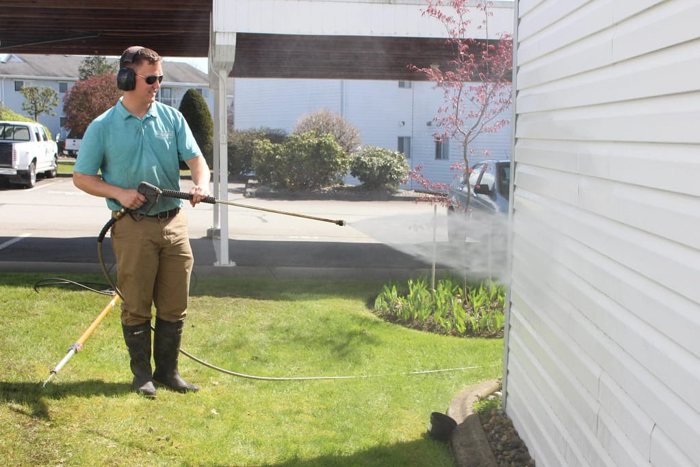 A man is wearing a light blue polo shirt while holding a pressure washer to clean the house.