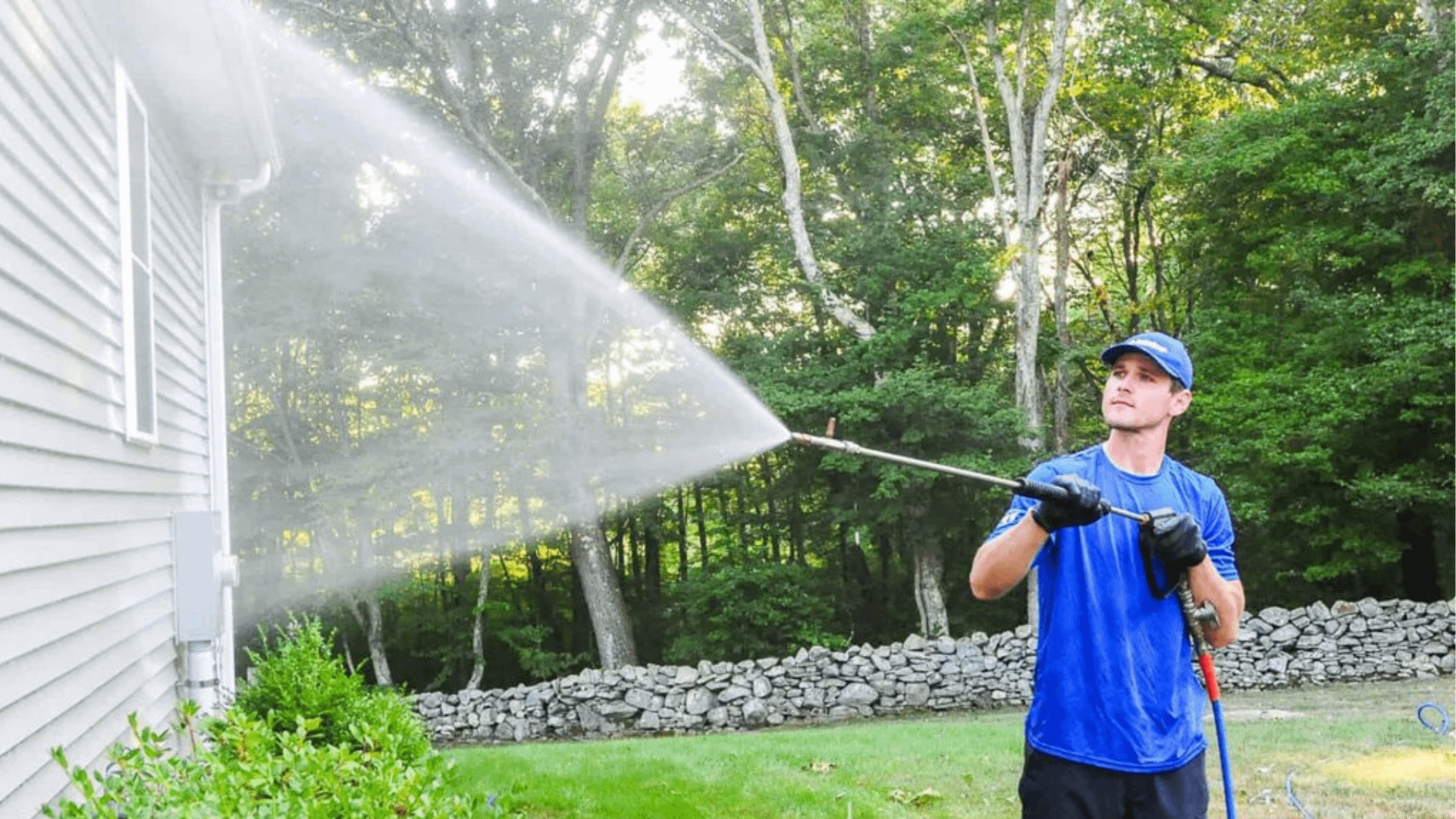 A man is wearing a hat and a blue t-shirt while softwashing a vinyl house as part of the house washing process