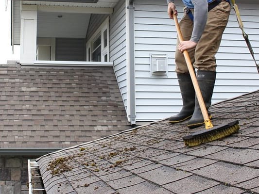 The man is holding a roofs brush to clean the roof while wearing his boots.