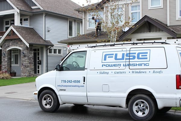 The fuse Power Washing Van is park the side of the residential area while the man is cleaning the top of the roof.